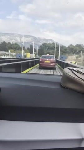 The video shows a car driving onto a floating bridge. The car is red and has a yellow taxi sign on top. The bridge is made of planks and has yellow barriers on the sides. The camera is inside a car, looking out through the windshield and side window. There is a purse on the dashboard. Through the windows, water and land can be seen. The floating bridge is a simple structure for vehicles to cross the water. The scene shows the car crossing from one side to the other. There is a knocking or banging sound.