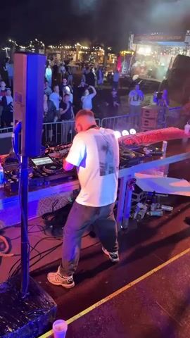 A DJ performs at an outdoor event at night. The DJ, wearing a white t-shirt and dark pants, is standing behind a DJ console with turntables and a mixer. Two large speakers are visible on the left side of the frame. A crowd of people stands behind a barrier in the background, some appearing to be watching the DJ. There are lights and tents in the background, suggesting a festival or market setting. Smoke machines are active, producing plumes of white smoke that rise into the air. A person in the foreground is partially visible, looking towards the DJ and holding a cup. The ground in front of the DJ booth is wet and reflective.