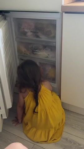 A young girl with long brown hair in a braid, wearing a yellow dress, sits on a light-colored floor, facing an open white freezer. The freezer has three transparent drawers, each labeled "FREEZEBOX" and filled with various items in plastic bags. The girl reaches her right hand into the bottom freezer drawer, appearing to search for something. The surrounding environment includes a white wall and a light wooden cabinet.
