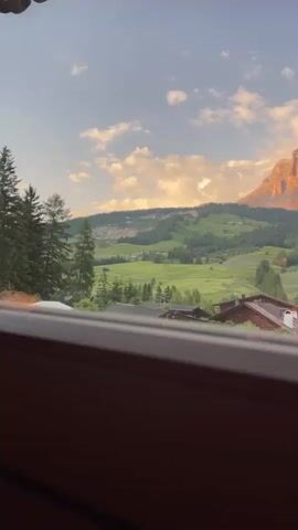 This video shows a scenic mountain landscape at sunset. The foreground is dominated by a wooden railing, implying the shot is taken from a balcony or window. Beyond the railing, a village with traditional wooden houses nestles in a valley. Lush green fields and scattered trees cover the rolling hills that lead up to a majestic mountain range. The mountain peaks are bathed in the warm, golden light of the setting sun, creating a dramatic and beautiful contrast with the shadowed valleys. The sky above is a blend of blue and soft pastel colors, with a few scattered clouds catching the sunlight. There is no visible text in the video. The overall mood is peaceful and serene, evoking the beauty of the Italian Dolomites during a tranquil evening. The location is identified as La Ila - Stern - La Villa, Italy.
