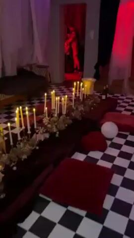 The video shows a room decorated with a black-and-white checkered floor. Along the floor are several burgundy cushions. A long table lined with candles, flowers, and a bottle of wine is on the left. Two doorways are visible, each with curtains. The first doorway has black and white curtains with red lighting. The second has a white curtain. People are standing in both doorways. The room is illuminated with red, blue, and purple lights around the ceiling. A bed and chair are on the left, and a white balloon is on the floor.