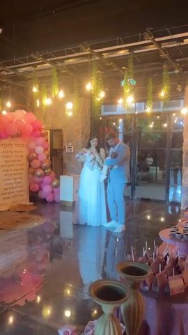 A couple stands in a well-lit indoor space, possibly a venue for an event. The woman, on the left, wears a long white dress and holds one baby. The man, on the right, wears a light blue suit and holds another baby. Both babies appear to be infants. Behind them, a wall is decorated with pink and gold balloons and a large scroll with text in Hebrew. Hanging from the ceiling are lights and greenery. The floor is dark and reflective, creating a mirrored effect. In the foreground, there are decorative objects, possibly part of a table setting. The overall atmosphere suggests a celebration, likely related to the birth of the babies.
