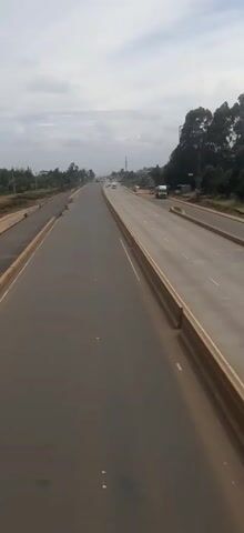 The video shows a wide, relatively empty highway under a cloudy sky. The road is divided, with one section appearing newer or recently repaved. A small truck is parked on the newer section. A person on a bicycle rides along this newer section, facing away from the camera. Trees line the edges of the highway, and a few cars and another truck are visible in the distance.