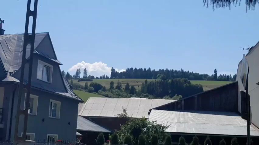 The video shows a rural landscape under a clear blue sky. In the foreground, a portion of a gray building with a gray roof is visible on the left, featuring a window. To the right, part of another building with a dark wooden exterior and a corrugated metal roof is present. In the middle ground, a large, light-colored corrugated metal roof dominates the scene, with a section of a darker structure partially obscuring it from view. Beyond these buildings, rolling green hills dotted with trees stretch into the background. A line of dark green coniferous trees forms a dense forest along the crest of the furthest hill, and a white cloud is visible in the sky above them. The overall impression is a sunny day in a serene, possibly agricultural, setting. There are no people or discernible actions taking place in the video.