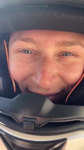 The video shows a person wearing a white motorcycle helmet with cat ears attached to it. Initially, the camera focuses closely on their face inside the helmet. The person smiles and has a nose ring. The camera then pulls back to reveal a scenic view behind them, showcasing a vast landscape of green hills, a town, and a body of water in the distance, all under a clear blue sky. The person's eyes are now more visible and appear blue. The camera angle shows a partial view of the person's black jacket. The camera continues to slightly adjust, showing more of the scenic view with the person's helmet still in the foreground.