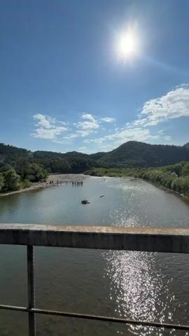 The video shows a wide river with calm, clear water reflecting the bright sun and blue sky. Lush green hills covered in trees surround the riverbanks. On the left bank, a group of people are gathered near the water's edge, some standing in the shallow river and others on the bank. A small, dark object, possibly a raft or debris, floats in the middle of the river. The view is framed by a metal railing in the foreground, suggesting the footage is taken from a bridge. The sun is high in the sky, casting a bright glare on the water.