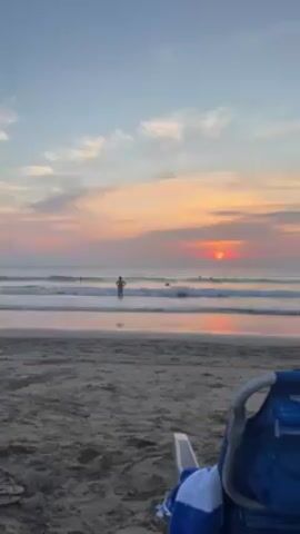 The scene is a beach at sunset. The sun, a vibrant orange, is low on the horizon, partially obscured by clouds. The sky is a mix of blue, pink, and orange hues. The water is calm, with small waves breaking on the shore. In the distance, a few people are wading in the water. In the foreground, there is a sandy beach and a blue beach chair with a blue and white towel draped over it. A pair of sandals are visible in the sand near the chair. The overall atmosphere is peaceful and serene.