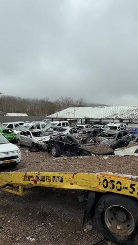 The video shows a car graveyard on a cloudy day. Many cars, wrecked and damaged, are piled together amidst dirt and debris. A yellow tow truck with Hebrew writing and the number 050-5531512 is parked nearby. The camera pans up to reveal the tow truck's crane mechanism. No people are visible in the video. The environment appears to be outdoors in an industrial or salvage yard setting.