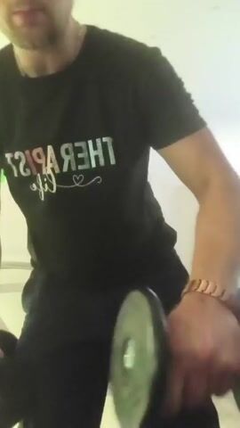 A person wearing a black t-shirt with writing on it and dark pants is performing dumbbell exercises in what appears to be a gym or workout space. The person is holding two dumbbells and is in motion, suggesting a series of repetitions. In the background, there is gym equipment including a treadmill, a dumbbell rack with weights, and an exercise ball. The lighting is bright, coming from overhead fixtures.