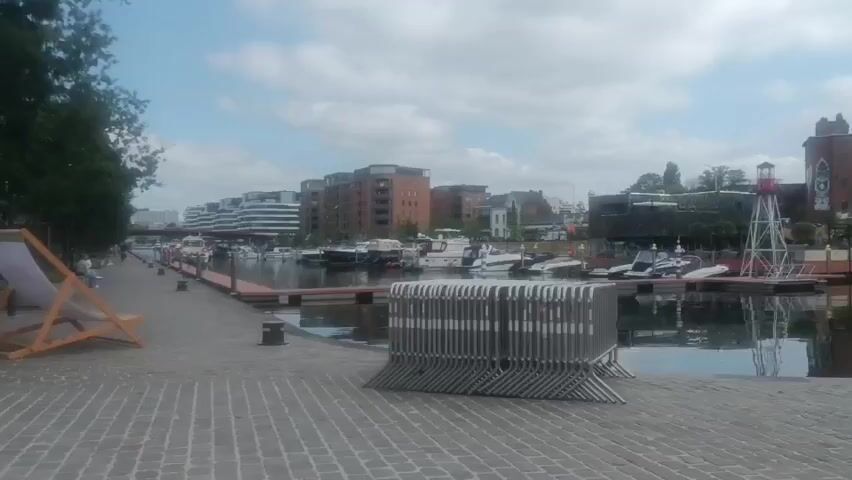 A wide shot captures a canal lined with boats and modern buildings. On the paved walkway, a folded stack of grey metal barriers stands in the foreground. To the left, a wooden deck chair with a white cushion sits near a tall, yellow sign. Several people are visible in the distance, walking along the canal. The sky is partly cloudy, with the sun illuminating the scene. A lighthouse-like structure is visible on the right side of the canal.