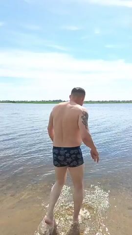 A man wearing dark-patterned swim trunks walks into a body of water. He is viewed from behind, and his back and shoulders are visible. The water is calm and appears shallow near the shore, gradually deepening as the man walks further in. The sky above is blue with scattered white clouds. The distant shoreline is lined with green foliage.
