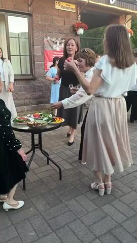 The video shows a group of people outdoors near a building with a sign that reads "РЕСТОРАН дом". In the foreground, a woman in a white top and a long beige skirt is standing, holding a glass. She is looking at another woman in a black top and gray skirt who is holding a small red object, possibly a strawberry, and a glass. This woman is leaning forward and appears to be offering the red object to the woman in white. There is a round table with a platter of food, including what looks like salads and fruit, between them. To the left, a woman in a dark green dress with white polka dots is visible, facing away from the camera. Further back, a woman in a white blazer and beige skirt stands with her arms outstretched. Another woman in a white top and skirt is visible in the background. The ground is paved with interlocking bricks. The lighting suggests it is daytime, possibly late afternoon or early evening. The overall atmosphere appears to be a social gathering or celebration.