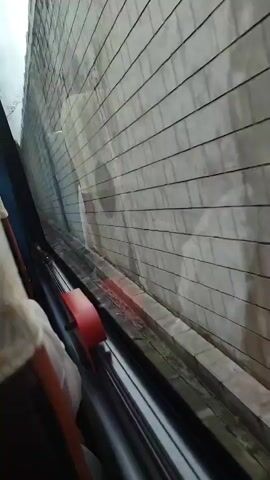 The video shows a view from inside a vehicle, looking out a window. The window is grimy and streaked, with what appears to be a concrete wall or structure just outside. The wall is made of large, rectangular tiles or blocks and has dark streaks, possibly from water damage or grime. In the foreground, part of a car interior is visible, including a seat and a red handle. The perspective suggests the vehicle is moving alongside the wall, creating a sense of motion. There are no other people or significant objects clearly visible besides the interior of the vehicle and the external wall.