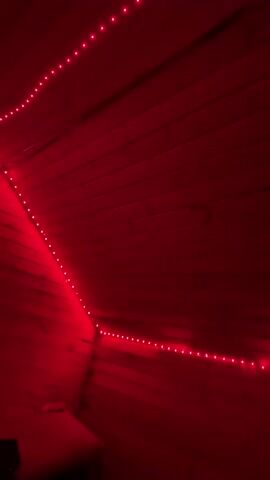 The video displays a close-up of a wooden paneled room illuminated by red LED strip lights. The lights are positioned along the seams where the wooden panels meet, forming a geometric pattern that emphasizes the angles of the room. The camera pans slowly across the textured wood grain, with the red glow creating a warm and immersive atmosphere. The scene is static, with no discernible people or actions. The focus remains on the interplay of light and shadow on the wooden surfaces.