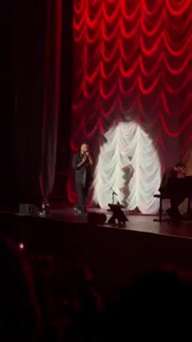 A male performer in a dark suit sings on a stage in front of a red and white draped curtain. The stage is illuminated by spotlights, and a pianist is visible to the performer's right. The audience sits in the dark, with some people holding up their phones, presumably to record the performance.