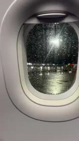 The video shows a close-up view from inside an airplane, looking out of a window. Rain is heavily falling on the window, obscuring the view outside. Through the rain-streaked window, blurry lights can be seen, suggesting a street or parking lot at night. The window frame is white and dominates the foreground. The scene is dark and wet.
