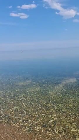 The video pans across a serene lakeside scene. The foreground is a rocky shore, with smooth, multi-colored pebbles of various sizes covering the beach and extending into the crystal-clear water. The water itself is incredibly transparent, allowing a clear view of the submerged rocks.

In the middle distance, the vast expanse of a calm lake stretches out, reflecting the pale blue sky. A few small, white clouds drift lazily overhead. The horizon is a soft, indistinct line where the water meets the sky.

In the far distance, a tiny, solitary figure can be seen on the water, possibly a boat or a person. This figure is so small it barely registers, emphasizing the immense scale and peaceful solitude of the location.

The time of day appears to be late morning or early afternoon, given the bright, diffused sunlight and the clear, blue sky with scattered clouds. The overall atmosphere is one of tranquility and natural beauty. The lack of any significant activity or human presence, beyond the distant figure, contributes to a feeling of quietude and vastness. The location, Bugul'deika, Russia, is known for its stunning natural landscapes, and this scene aligns with that description, showcasing the pristine beauty of a Russian lake. There is no visible text in the media file.