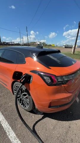 A bright orange electric car is parked on a gravel lot, its charging port open and a black charging cable plugged in. The car has a black roof and tinted windows. The background shows a clear blue sky with scattered white clouds, power lines, and some distant buildings. The overall scene depicts a car being charged outdoors on a sunny day.