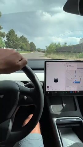 The video shows the interior of a car, likely a Tesla, from the perspective of the driver. The driver's left hand is visible gripping the steering wheel. The car is moving on a paved road lined with trees and houses. The car's large central touchscreen display is visible, showing a navigation map with a blue route highlighted, indicating the car is following directions. The time on the display changes from 23 to 49. The sky is cloudy.
