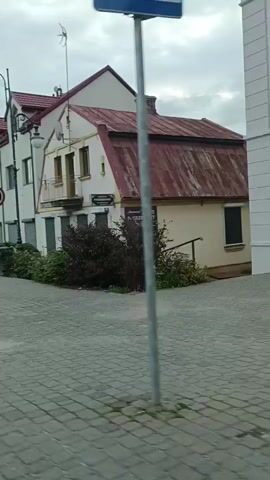 The video shows a view from a moving vehicle on a cobblestone street. The camera pans to the right, revealing a building with a red, corrugated metal roof and white walls. The building has multiple windows, some with closed shutters. There is a street lamp in front of the building, and a speed limit sign showing 40 is visible. A dark-colored car is parked on the street. Further down the street, another building with a similar architectural style is visible, also with a red roof and white walls. A white Audi station wagon with a license plate is driving away from the camera. The sky is overcast. There are bushes and some greenery in front of the buildings. The overall impression is a quiet, possibly European, town or village setting.