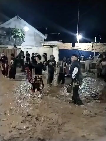 At night, an outdoor scene in Jenggrik, Indonesia, shows several individuals engaged in a performance or ritual in a muddy, unpaved area. The ground is covered in mud and puddles, suggesting recent rain. Artificial lights illuminate the scene, casting shadows.

Most people are men, dressed in various outfits; some wear black tops and pants, while others incorporate traditional elements. In the center, a person in patterned shorts and a black top is actively moving, possibly dancing, with splashes of mud around them. On the right, another individual carries a large prop, possibly a part of a traditional costume or a stylized animal figure, made of white and dark material. Other participants are moving around, some appear to be watching, while others are holding props or instruments, such as a drum visible on the left.

In the background, a white building and some wooden structures are visible, along with a blue tarp, suggesting a community or village setting. The atmosphere appears energetic and celebratory, indicative of a cultural event or parade. No discernible text is visible on clothing or objects.