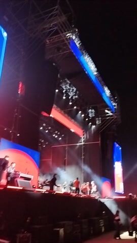 The video captures a lively nighttime concert scene in Maipú, Chile. The primary subjects are a large, enthusiastic crowd and intricate stage structures. Elevated platforms and ramps, brightly illuminated with red lights, are visible, suggesting a dynamic performance area. Numerous spotlights cut through the dark sky, creating beams of white light and illuminating clouds of smoke or fog emanating from the stage.

The camera pans across the scene, showcasing the dense gathering of people who are enjoying the event, bathed in the vibrant red glow of the stage lighting. The overall atmosphere is energetic and festive, typical of a large-scale music event. No discernible text is visible in the frames.