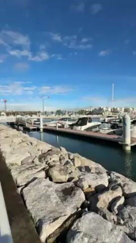 The video shows a marina on a sunny day with a partly cloudy blue sky. The camera is positioned behind a rocky breakwater, looking out over a harbor filled with boats docked at floating piers. The boats vary in size and type, from smaller motorboats to larger yachts, some with sails. In the background, there are white buildings, potentially apartments or commercial properties, lining the harbor. The water in the marina is calm and reflective, and the overall scene conveys a sense of tranquility and leisure. There are no people visible in the frame.