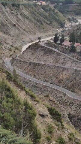 A drone shot looking down on a winding road carved into a steep, dry, mountainous terrain. The road has several sharp switchbacks. A river flows at the bottom of the valley, and a few small buildings are visible on the slopes and in the valley. The landscape is rocky and sparse, with some green vegetation on the lower slopes. There are a few cars visible on the road. The sky is overcast.