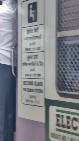 This is a video taken from a train platform. Several people are standing in the open doorway of a train carriage, with some hanging out of the windows. The train appears to be at a station, as there is a platform with tactile paving visible. A person in a white shirt and dark pants is leaning out of the train doorway. The side of the train has advertisements, including one for ELECTRAL ORS. The video pans along the side of the train, showing more people hanging out of the windows and doorways. The environment appears to be an urban train station, possibly in India, given the text in Hindi visible on the train.