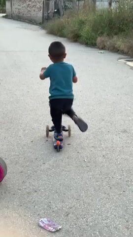 A young boy is seen from behind, riding a three-wheeled scooter on a paved street or alley in Karadjovo, Bulgaria. He is wearing a teal t-shirt and dark pants, propelling the blue and orange scooter with his right foot. An adult male, dressed in a black t-shirt and shorts, walks in the background behind the boy.

The setting is an outdoor residential area with buildings and a brick wall visible on the right side of the frame. The ground is paved with a light gray surface, possibly asphalt or concrete. On the left, there's some overgrown vegetation and a partial view of another pink toy in the immediate foreground. The lighting suggests it is daytime, likely a clear and sunny day. The scene depicts a casual moment of outdoor play. No visible text is clearly legible within the frame.