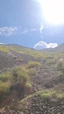 The camera pans across a rocky, hilly landscape under a blue sky with some white clouds. The ground is covered in a mix of small rocks and sparse green vegetation. In the distance, a larger hill rises, also covered in rocks and patches of green. The sun is shining brightly, causing some glare in the video. The overall scene suggests a rugged, natural environment.