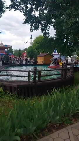 A wide shot shows an amusement park on a cloudy day. In the foreground, there is a large tree with green leaves and a wooden fence surrounding a circular pond. Several swan-shaped boats are in the pond, with people riding in them. In the background, there are various amusement park rides, including a carousel and a Ferris wheel. People are walking around the park, and a mosque-like building with a tall minaret can be seen in the distance. The overall atmosphere appears to be lively and cheerful.