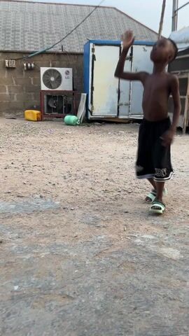 A young, dark-skinned boy, shirtless and wearing black shorts and light-colored sandals, is playing with a white soccer ball on a dirt and concrete surface. He dribbles the ball with his bare feet and the camera follows the action closely. In the background, there is a brick wall, a white building with a blue door, an LG air conditioning unit, and general debris. The sky is overcast, suggesting it might be early morning or late afternoon. The overall setting appears to be a residential or semi-industrial yard.