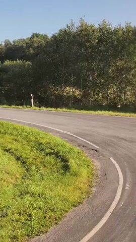 A gray station wagon drives around a bend in a rural road on a sunny day. The road is paved and bordered by green grass and trees. The car is the only visible object on the road.