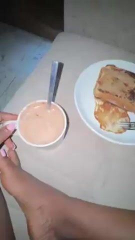 The video shows a breakfast of toast with a spread, an egg omelet, and a beverage in a cup with a spoon. The food is on a white plate, placed on a beige patterned surface with a visible foot in the lower left corner. The floor is visible in the corner, with light-colored tiles.