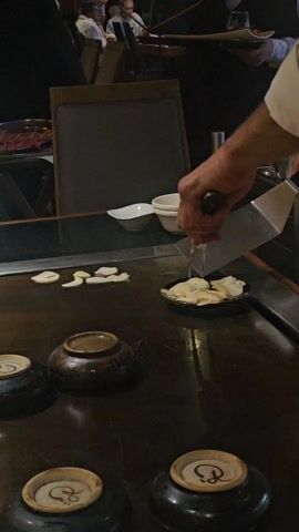 The video shows a chef at a hibachi grill preparing food in a restaurant. The chef, wearing a white shirt, uses a spatula and tongs to move slices of garlic on the hot grill surface. Some garlic is already in a small black dish. There are several small ceramic bowls arranged on the grill around the cooking area. In the background, other diners can be seen seated at tables and a waiter carries a tray of food. The setting is a dimly lit restaurant.