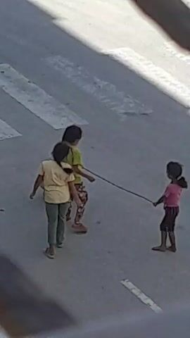 The video shows three young children playing a game with a rope on a paved surface. A person wearing a light-colored shirt and dark pants is partially visible on the left, possibly kneeling or bending down. Two girls are standing in the center, facing away from the camera. One girl wears a yellow t-shirt and green pants, with her hair in a ponytail. The other girl wears a lime green t-shirt and patterned pants. A third child, wearing a pink t-shirt and striped shorts, is on the right, holding one end of a dark rope. The girl in the green t-shirt is holding the other end of the rope. The children appear to be engaged in a tug-of-war or a similar game, as the rope is taut between them. The background consists of a grey, textured surface with some lighter patches. The lighting suggests it is daytime. The video is grainy and the resolution is low, making it difficult to discern fine details.