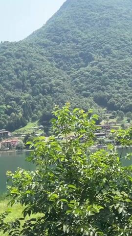 A directional sign indicating a left turn is visible in the foreground, positioned in front of a scenic landscape. Behind the sign, a body of water, likely a lake, stretches out. Along the far shore of the lake, a small town or village with houses is nestled against a backdrop of a densely forested mountain. The sky is clear and the overall scene suggests a bright, sunny day in a natural environment. The camera movement appears to be a slow pan or slight drift to the right, revealing more of the lakeside setting.