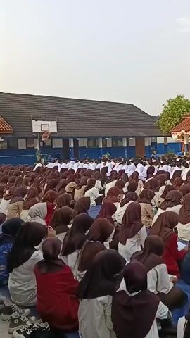 A large group of students, mostly female and wearing hijabs in various shades of brown, white, and maroon, are sitting on the ground in rows in an outdoor courtyard. They are facing a building with a red-tiled roof and blue trim, where a banner is displayed with text and images. A flagpole with a red and white flag is visible to the right of the banner. The students appear to be attentive, with some holding books or papers. The sky is overcast.