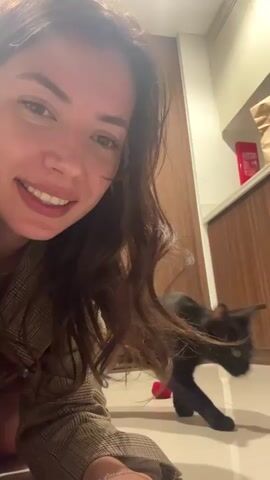 A woman is holding a camera and recording herself and a black cat. The cat is initially walking around on a tile floor near kitchen cabinets. The cat then rolls onto its back and looks at the camera. The woman smiles and lowers the camera so that her face is closer to the screen. The cat is still lying on its back.