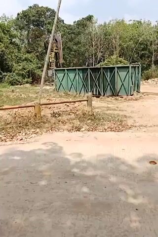 The video shows an outdoor scene with a dirt road in the foreground. Behind the road, there is a closed gate with a rusty horizontal bar. To the left of the gate, a tall, slender pole extends upwards, partially obscuring the view. Further back, an oil pumpjack is visible against a backdrop of lush green trees. Adjacent to the oil pumpjack is a large, green, rectangular storage tank, divided into sections by crossbeams. The ground around the gate and tank is covered in dry leaves and debris. The sky is partly cloudy. The scene appears to be a rural or industrial area, possibly an oil extraction site.