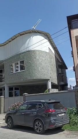 The video shows a street scene with a modern building on the right and other buildings and trees on the left. A dark SUV is parked in front of the building. The building features a curved roof and a stone facade. The sky is blue with some clouds. The camera pans slowly to the left, revealing more of the street and the surrounding environment, including a lush green hillside in the background.