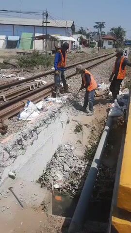 Three construction workers are seen working on railroad tracks under a clear sky. The worker on the left is operating a jackhammer, dressed in a blue long-sleeved shirt, orange vest, and black boots. The middle worker, wearing a hat, blue long-sleeved shirt, and orange vest, is using a metal rod to break apart concrete. The worker on the right, wearing an orange vest and black pants, stands by, observing. The area is littered with debris, including torn bags, concrete chunks, and rebar, suggesting an ongoing demolition or construction project. A yellow platform is visible on the right edge of the frame, and railroad tracks extend into the distance. Several buildings and vegetation are visible in the background.