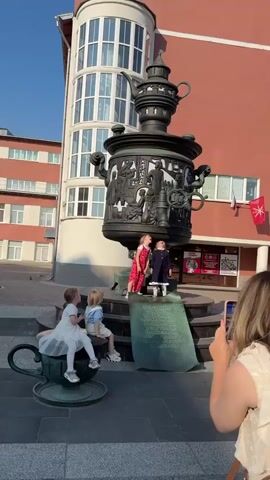 The video opens with a wide shot of a large, ornate samovar sculpture in what appears to be a public square. Several young children are interacting with the sculpture. In the foreground, a woman holds up a mobile phone, seemingly filming or taking pictures. To the left, two children, a boy and a girl, are seated on a large teacup sculpture, which is part of the overall installation. The girl on the teacup is wearing a white dress. The boy is wearing a light blue shirt and shorts.

In the center of the frame, on a lower platform of the samovar sculpture, stand two young girls. The girl on the left wears a red and white patterned jumpsuit. The girl on the right wears a dark blue dress with white trim and ruffled socks. Both are looking upwards towards the samovar.

Behind the central group, another young boy is visible, wearing a green t-shirt, looking towards the main group. The background features a building with a distinctive curved, multi-story facade and red brick walls, interspersed with windows. There are some signs on the building. The lighting suggests it is daytime, with bright sunlight. The ground is paved. The overall atmosphere is one of playful exploration of a public art installation by children.