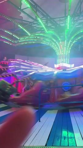 The video shows a carnival ride with flashing lights and spinning cars. The ride appears to be a spinning teacup-style attraction with individual cars that rotate. The lights are a mixture of green, pink, and blue, creating a festive atmosphere. The camera is held by a person, and their thumb is visible in the foreground. The ride is in motion, with the cars blurred due to the fast spinning. The background shows what appears to be a tent or covered area of a carnival.