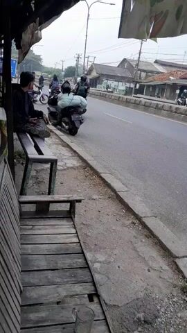 A person is sitting on a wooden bench. In the background, there is a road with traffic. Several motorcycles are visible, one of which is carrying a large package on the back. A truck is also driving on the road. The scene appears to be in a roadside area with some buildings and vegetation.