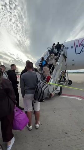 This is a low-angle, vertical video taken outdoors at an airport. The focus is on a group of people boarding an airplane via a mobile staircase. The plane, identified by the "WIZZ" logo in large purple letters on its side, is white with pink accents. Several passengers are visible ascending the metal stairs, some carrying luggage. In the foreground, three people are waiting their turn to board. One person in a grey shirt and white shorts faces away from the camera, a duffel bag slung over their shoulder. Another person in a black hoodie is partially visible on the left. The ground is paved tarmac, marked with red and white lines. The sky above is cloudy with patches of blue. The video pans slightly to the right, showing more of the plane's fuselage and a pink engine nacelle.