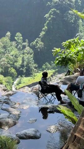 Two people are sitting on camping chairs at the edge of a rocky waterfall overlooking a lush green valley. One person, wearing a dark patterned shirt and pants, is facing away from the camera, looking out at the view. The other person, partially visible, is also facing away from the camera, wearing a white shirt and dark shorts. The environment is a jungle with dense trees and a waterfall cascading down a cliff face. In the distance, a volcano emerges from the clouds. The water in the foreground reflects the sky and surrounding foliage. The scene is bathed in natural daylight.