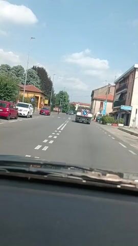 The video shows a view from inside a car, looking out through the windshield. The car is driving down a paved road in what appears to be a European town. The road is lined with buildings and trees. Several cars are visible on the road, including a white SUV, a red sedan, a dark-colored sedan, and a truck. There is also a person on a bicycle in the distance. The sky is blue with some clouds. The video is 4 seconds long.