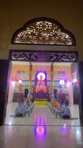 The video shows an interior shot of a Buddhist temple. In the center of the frame, a golden statue of Buddha is illuminated by a bright light behind it. In front of the statue, there are several lit candles. The walls of the temple are decorated with paintings of landscapes and ornate carvings. In the foreground, two rows of people are seated on the floor, facing the statue. The people are dressed in light-colored robes, suggesting they are monks or nuns. They appear to be engaged in prayer or meditation. The lighting in the temple is dim, with colorful lights reflecting on the polished floor. The overall atmosphere is one of solemnity and reverence.